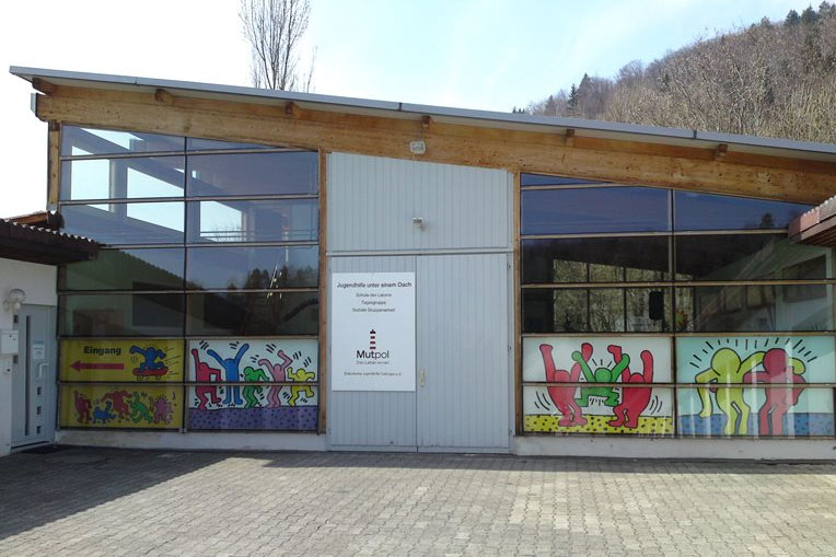 Gebäudefront mit großen Fenstern, bunten Wandmalereien und einem Schild, das auf eine Jugendveranstaltung unter einem Dach hinweist.