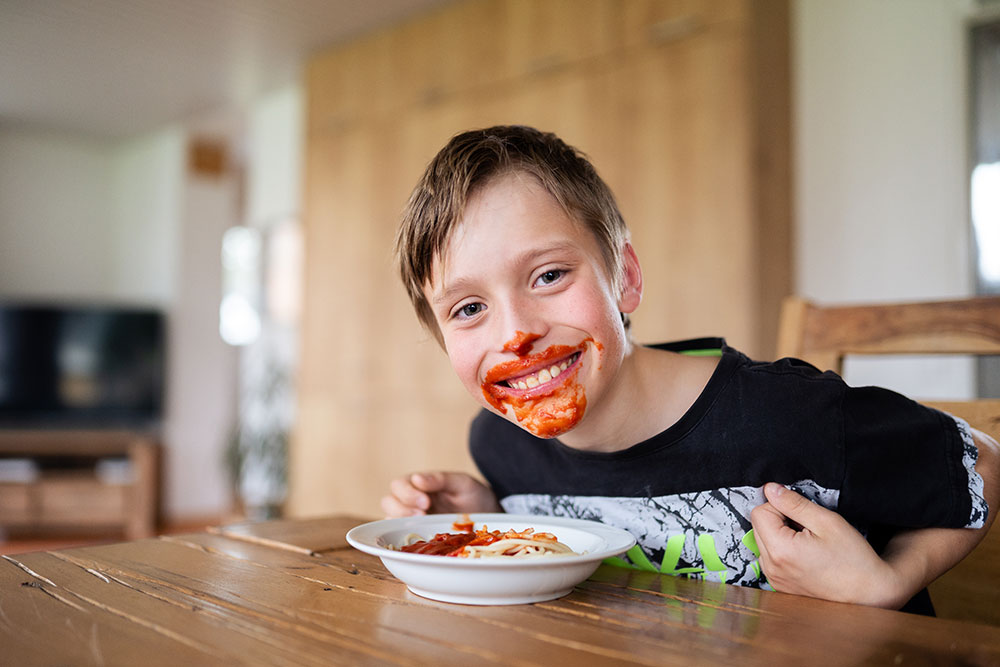 Junge mit verschmiertem Tomatensoße im Gesicht, lächelnd beim Essen von Spaghetti am Esstisch.