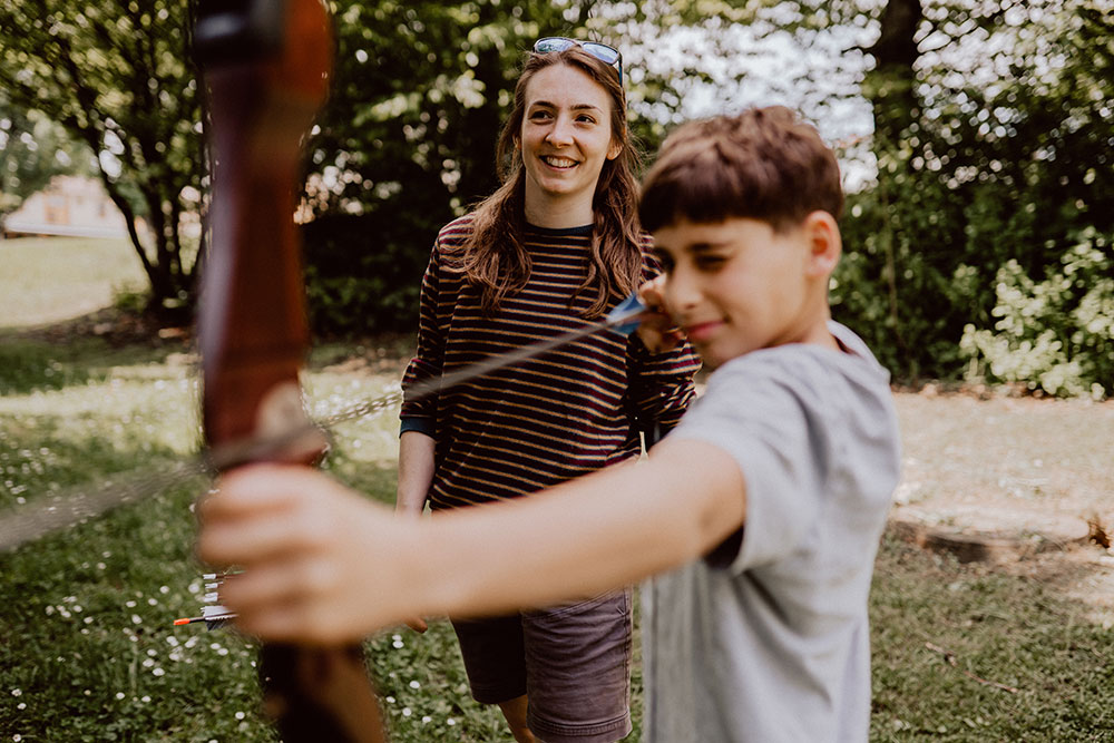 Ein Junge schießt mit einem Bogen auf eine Zielscheibe, während eine lachende Frau im Hintergrund im Garten zuschaut.