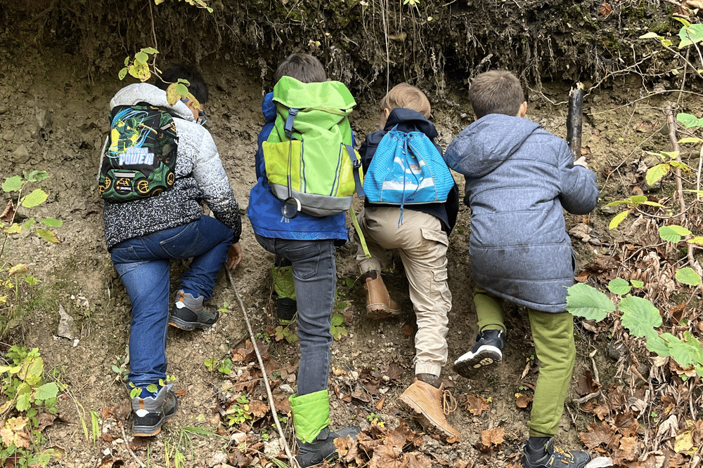 Vier Kinder mit Rucksäcken klettern an einer steilen, erdigen Steigung im Wald nach oben.