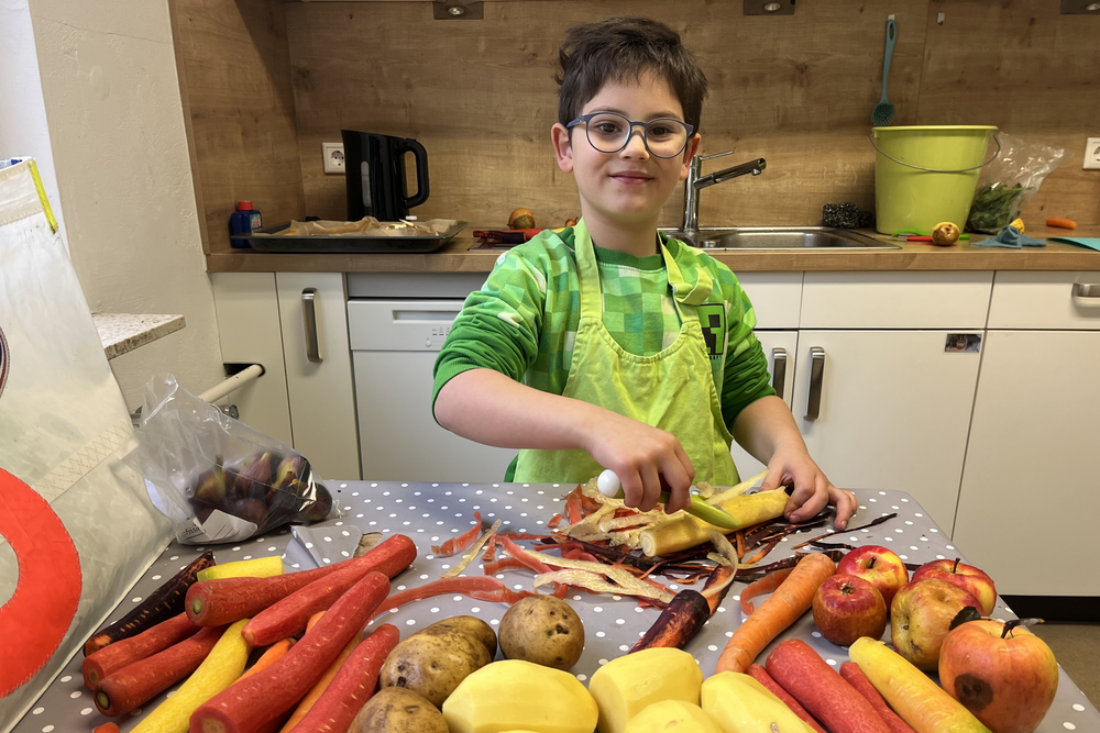 Junge mit Brille und grünem Hemd beim Schälen und Zubereiten von Gemüse in der Küche, umgeben von Karotten, Äpfeln, Kartoffeln und anderem Obst und Gemüse.
