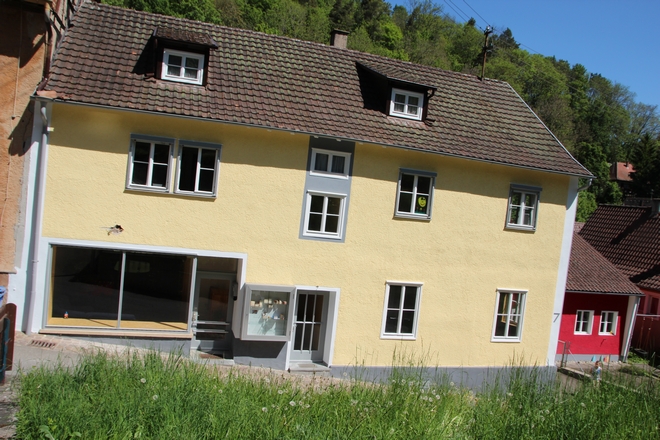 Mehrstöckiges Haus mit gelber Fassade, weißen Fenstern und Dachgauben in einer ländlichen Umgebung.