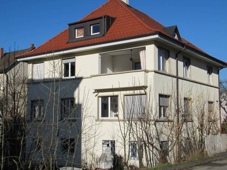 Mehrfamilienhaus mit weißer Fassade, roten Dach und mehreren Balkonen, umgeben von Bäumen ohne Blätter.
