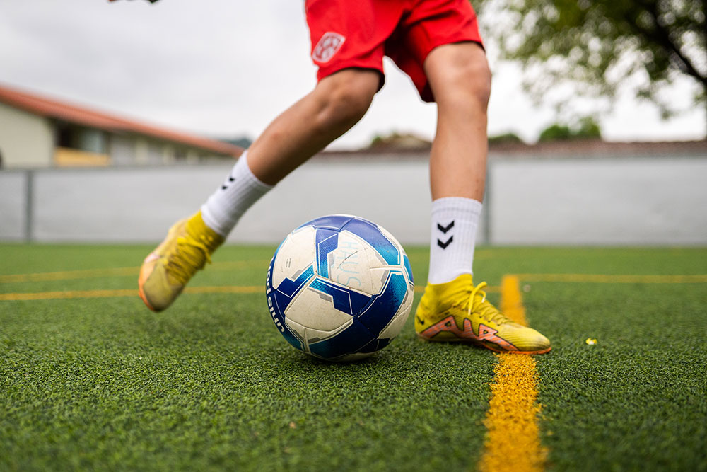 Fußballspieler in roten Shorts und gelben Schuhen beim Dribbling auf einem grünen Fußballfeld.