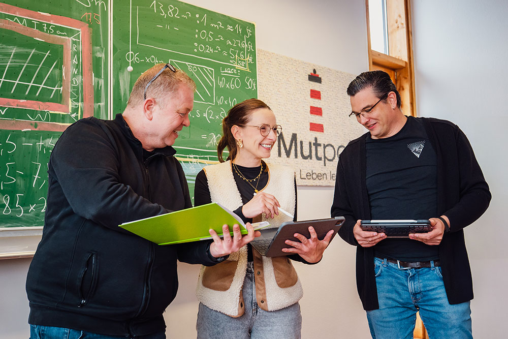 Drei Erwachsene stehen in einem Klassenzimmer vor einer grünen Tafel mit mathematischen Formeln und lachen, während sie mit Tablets arbeiten.