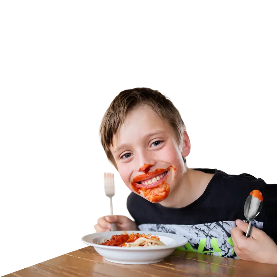 Junge mit Tomatensoße im Gesicht, isst Spaghetti und hält eine Gabel und einen Löffel, während er lächelnd in die Kamera schaut.