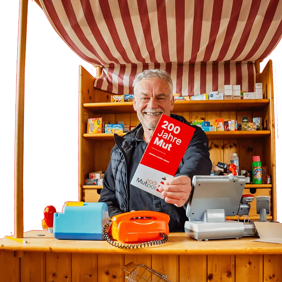 Ein älterer Mann steht an einem Holzverkaufsstand, hält ein rotes und weißes Schild mit der Aufschrift "200 Jahre Mut" und lächelt.