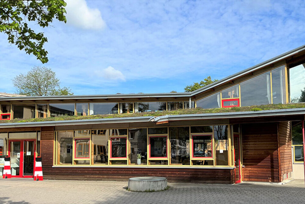 Modernes Gebäude mit großen Fenstern, Holzfassade und begrüntem Dach unter blauem Himmel.