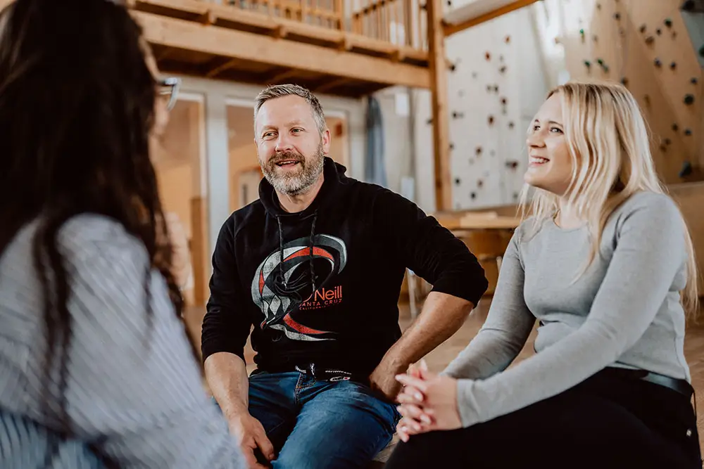 Drei Menschen sitzen im Gespräch in einem gemütlichen Raum mit Holzdecke und Kletterwand im Hintergrund.