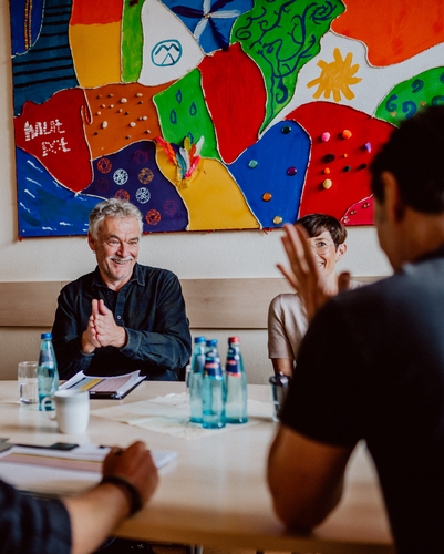 Drei Menschen sitzen in einer Besprechung vor einem bunten, abstrakten Wandbild, und einer von ihnen klatscht.