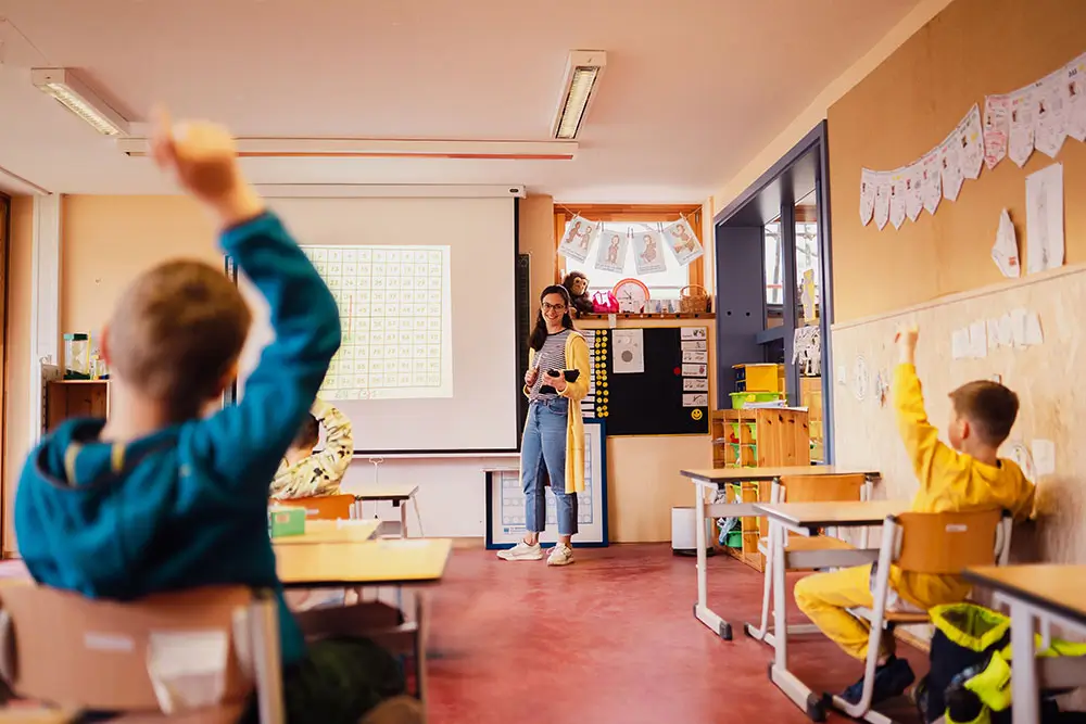 Eine Lehrerin steht vor einer Tafel in einem Klassenraum, während Kinder im Unterricht sitzen und eine Hand heben.