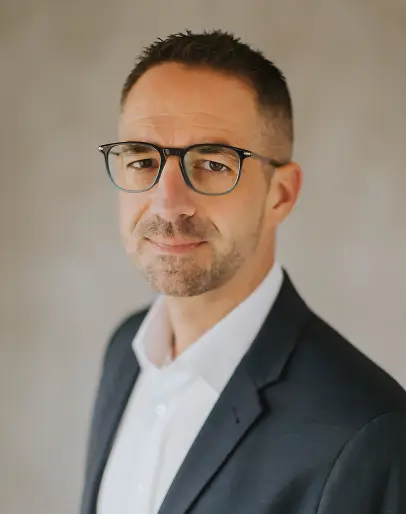 Porträt eines Mannes im Anzug mit Brille vor hellem Hintergrund.