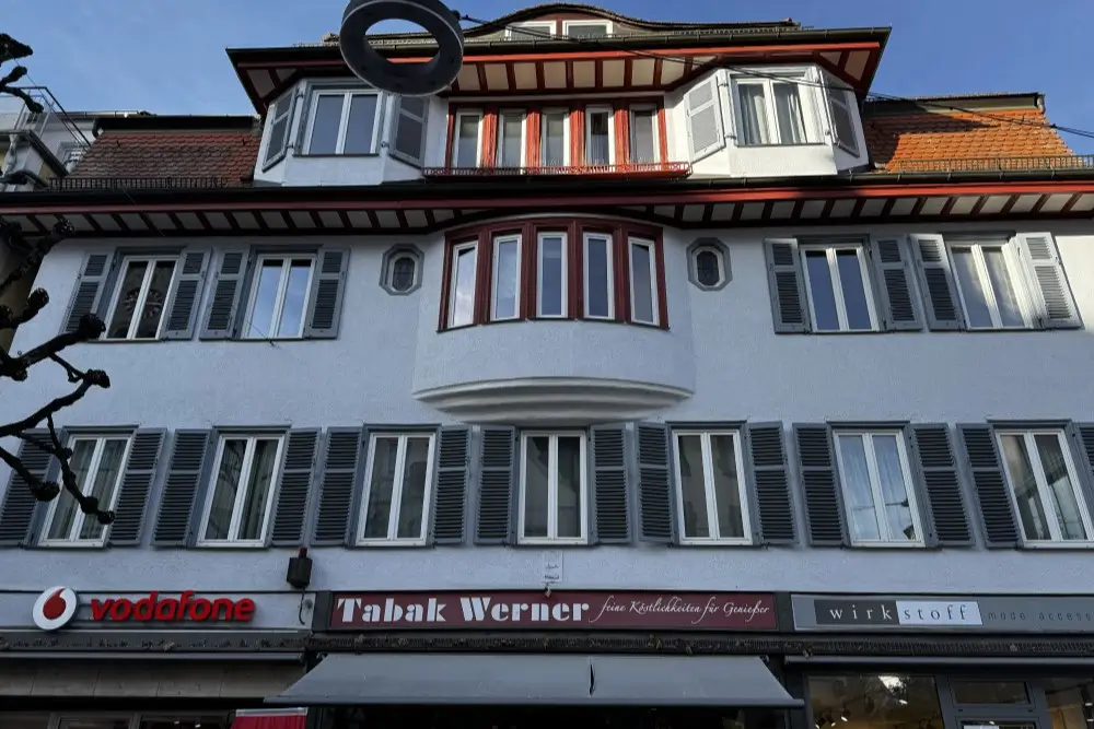 Weißes Mehrfamilienhaus mit grauen Fensterläden; Erdgeschoss mit Vodafone-Logo.