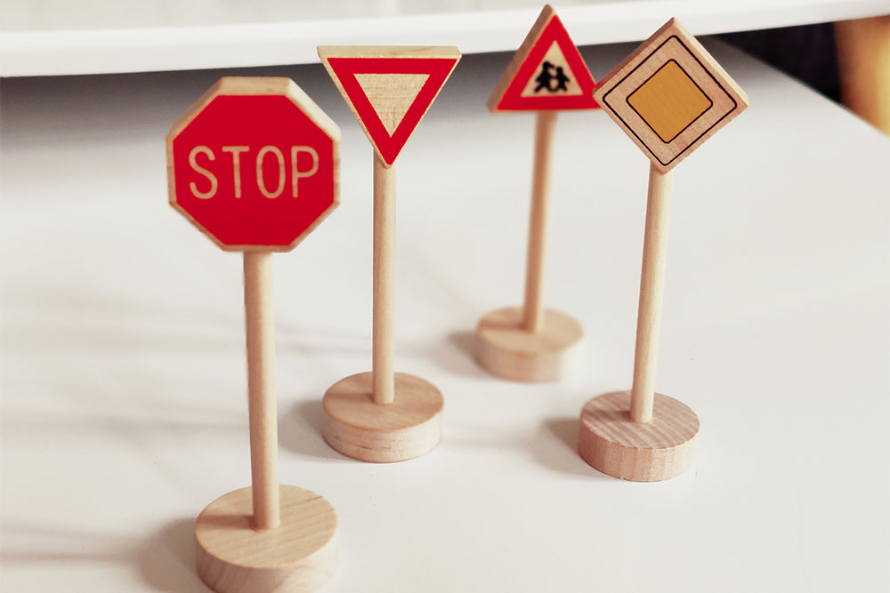 Vier kleine Holz-Straßenschilder auf Holzstäbchen, darunter ein Stoppschild, ein Vorsicht-Schild, ein Schulweg-Schild und ein Vorfahrtszeichen.