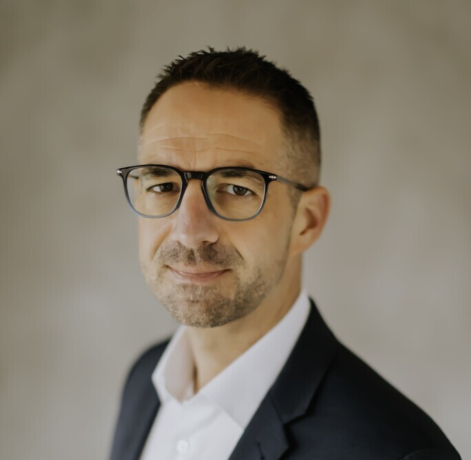 Porträt eines Mannes in Brille und Anzug vor neutralem Hintergrund.