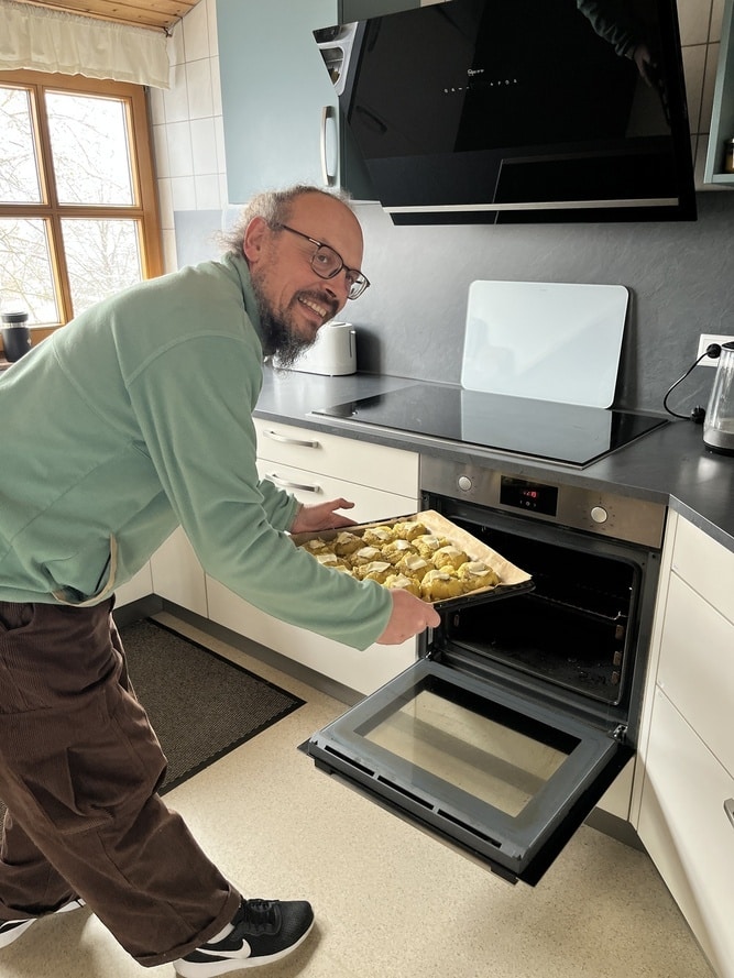 Mann mit Brille und Bart schiebt Backblech mit Kartoffelstücken in den Ofen.